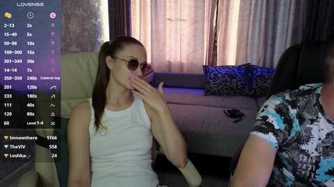 Video of a light-skinned woman with long brown hair, wearing sunglasses and a white dress, seated in a dimly-lit living room with a gray couch and patterned pillows.