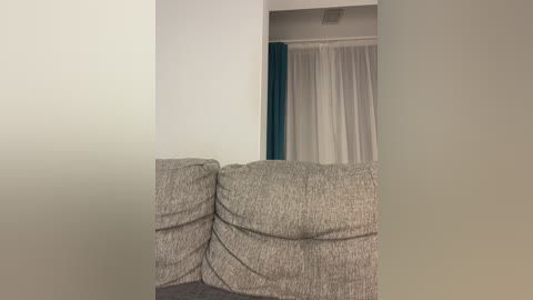 Video of a minimalist living room featuring a beige sectional sofa with textured fabric, a white curtain, and a teal accent wall, creating a serene and modern atmosphere.