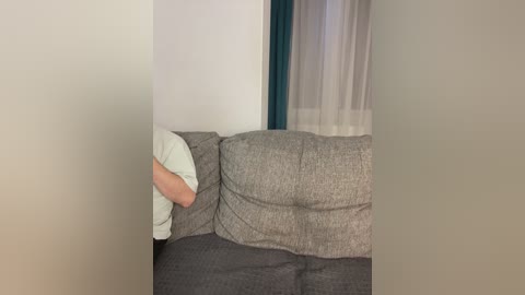 Video of a person, partially obscured by a blurry white curtain, resting their head on a gray fabric sofa with a dark cushion. The background includes a white wall and a teal curtain.