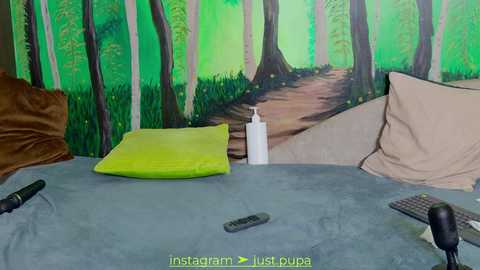 Media: A video of a bedroom with a green forest mural on the wall, featuring a tree-lined path. The bed has a blue-gray duvet, green pillow, beige pillows, and a white water bottle.