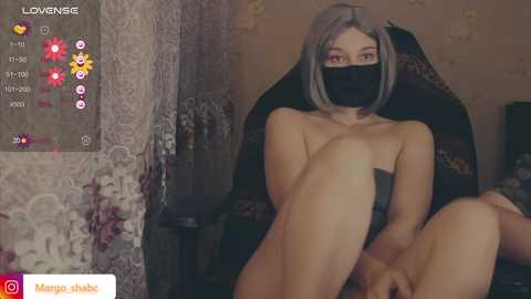A video of a topless Asian woman with short, straight grey hair, wearing a black face mask, sitting in a dark gaming chair, in a dimly lit room with floral wallpaper and a gaming overlay.