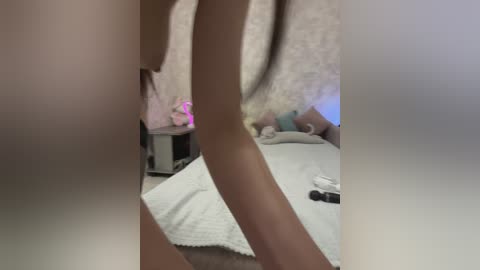 A blurry, candid video captures a naked person with light skin, possibly female, in a dimly lit bedroom. The background features a bed with pillows, a toy, and a furry blanket.