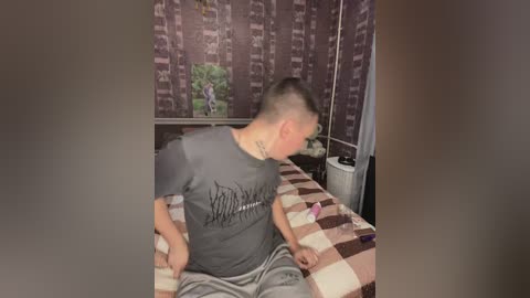 Media: Video of a man in a gray t-shirt and gray shorts, sitting on a bed with a brown and beige checkered blanket. The room features dark purple wallpaper with a tribal pattern and a window with a green plant.
