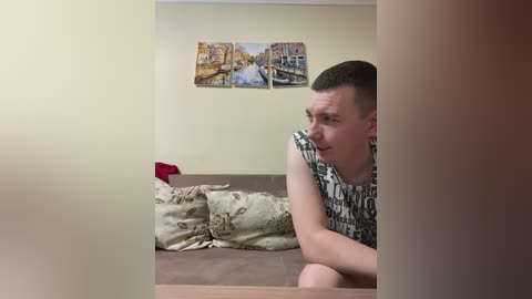 Media: Video of a young man with short brown hair, wearing a black and white graphic t-shirt, lying on a beige couch with patterned pillows, gazing at a painting of a canal scene on a cream wall.