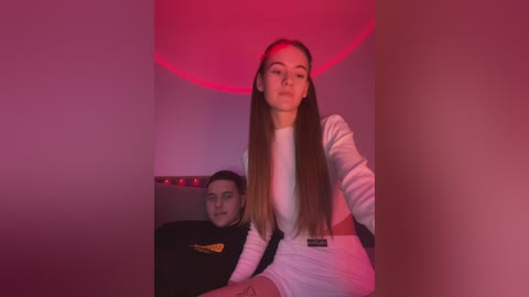 Media: Video of a young woman with long, straight brown hair, wearing a white top, standing in front of a man with glasses, both under a red light, in a dimly lit room.