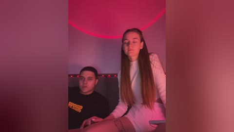 Media: Video of a young man and woman in dimly lit, red-hued room; man in black shirt, woman in white top and shorts, both with long hair, sitting close.