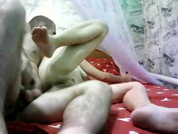 Video of a naked man and woman engaged in intimate position on a red bed with white hearts, inside a tent-like room with textured, greyish walls.