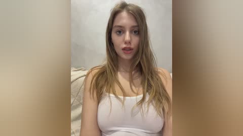 Media: Video of a young Caucasian woman with long, straight blonde hair, wearing a white tank top, sitting on a beige couch. Her expression is neutral, and the background is a plain, light-colored wall.