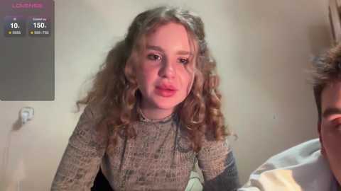 Media: Video of a young woman with curly brown hair, wearing a gray sweater, in a beige room, partially visible man in background, with a video game screen showing 10/150 points.