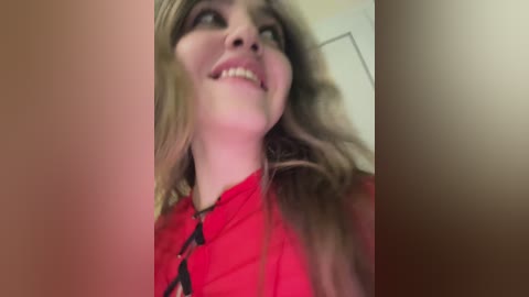 Media: Video of a young Caucasian woman with long, wavy light brown hair, wearing a red top, smiling broadly, with a slightly out-of-focus background.