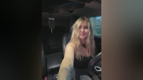 A video of a blonde woman with shoulder-length hair, wearing a black tank top, sitting in the driver's seat of a car. The car's interior is dimly lit, with black leather seats and a steering wheel.
