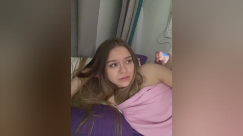 Video of a young woman with long, wavy brown hair, lying on a purple bedspread, wearing a pink top, looking thoughtful, surrounded by gray and white curtains in a dimly lit room.