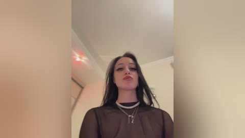A video of a young woman with long black hair, wearing a sheer black top and silver necklaces, standing in a beige hallway with a blurred red light source in the background.