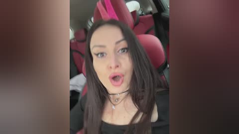 Media: A video of a young woman with long, dark hair and pale skin, wearing a black top and a choker necklace, sitting in a red car seat. She has a surprised expression.