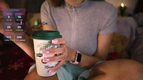 Media: A video showing a woman in a grey shirt, holding a coffee cup with a green lid, sitting on a plush couch with a teddy bear. A virtual overlay displays health metrics.
