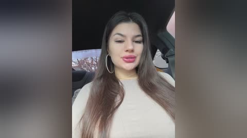 Media: Video of a young woman with long, straight dark hair, fair skin, and a curvy figure, wearing a beige top and large silver hoop earrings. She has a neutral expression, sitting in a car with a blurred background.