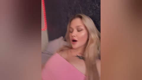 Media: Video of a blonde woman with fair skin, medium breasts, and a tattoo on her chest, lying on a bed, eyes closed, mouth open in apparent pleasure, surrounded by red lighting.