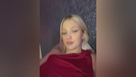 Media: Video of a fair-skinned, blonde woman with closed eyes, wearing a red shirt, lying on a dark textured surface.