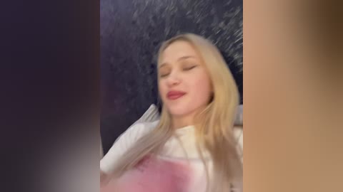 Media: Video of a blonde woman with closed eyes, wearing a white shirt, against a textured, dark background, creating a moody, intimate atmosphere.