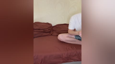 Media: A video of a person, possibly a man, sitting cross-legged on a bed with maroon sheets, wearing a white T-shirt and shorts. The background features a cream-colored wall with a subtle floral pattern.