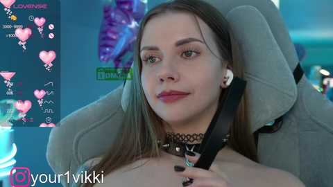 Media: Video of a fair-skinned, young woman with straight, long brown hair, wearing headphones, a black choker, and a black leather seatbelt across her chest. Background features a blue light and a screen displaying chat icons and hearts.