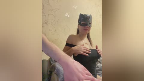 Video of a woman in a black cat mask and black leather outfit, holding her breasts, in a dimly lit room with floral wallpaper and a grey blanket.