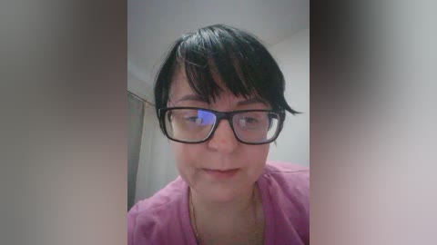 Media: Video of a short-haired woman with glasses, wearing a pink top, standing indoors with blurred background.