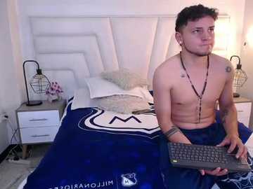 Video of a shirtless, lean, young man with short dark hair, tattoos, and a necklace, sitting on a bed with a blue duvet, holding a keyboard, in a minimalist bedroom with white furniture.