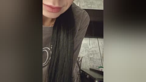 Video of a woman with long, straight black hair, wearing a grey graphic tee and cardigan, standing in a dimly lit room with a brick wall and dark furniture.
