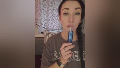 Video of a young woman with long dark hair, wearing a gray \"AUBURN\" t-shirt, blowing into a blue inhaler, in a room with patterned wallpaper and a wooden table.