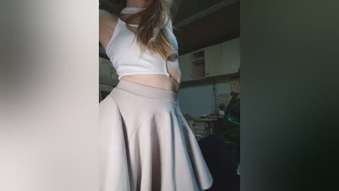 Video of a young woman with long, blonde hair wearing a white crop top and a high-waisted, light grey skirt, standing in a dimly lit room with shelves and clutter in the background.