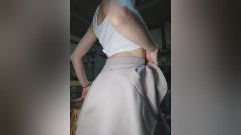 Video of a slender, fair-skinned woman with light brown hair, wearing a white tank top and high-waisted beige skirt, standing in a dimly lit room with wooden furniture.