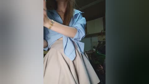 Video of a woman with long brown hair, wearing a light blue button-up shirt tied at the waist, exposing a high-waisted beige skirt, standing in a dimly lit room with kitchen cabinets in the background.