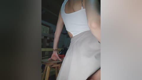 Video of a woman's back in a white tank top and high-waisted gray skirt, leaning on a wooden stool in a dimly lit room with exposed brick walls and scattered objects.