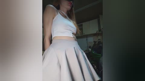 A video of a slender, light-skinned woman with long, straight brown hair, wearing a white tank top and high-waisted, pleated skirt, standing in a dimly lit room with a cluttered background.
