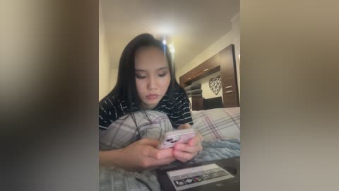 Video of an Asian woman with long black hair, wearing a black and white striped shirt, lying on a bed reading a magazine, in a dimly lit room with beige walls and a wooden headboard.