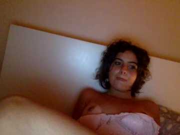 Video of a young woman with curly black hair, fair skin, and small breasts, wearing a light pink bra, lying on a beige bed in a dimly lit room.