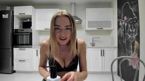 Media: Video of a blonde woman in a black lace bra, sitting at a kitchen table with a black coffee cup, in a modern, white kitchen with black appliances and a chalkboard wall.