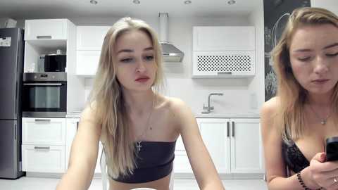 Media: Video of a young blonde woman with fair skin, wearing a black strapless top, sitting in a modern kitchen with white cabinets and stainless steel appliances.