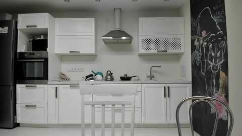 Media: Video of a minimalist white kitchen with sleek cabinets, stainless steel appliances, a white island with three stools, and a blackboard wall with chalk drawings.