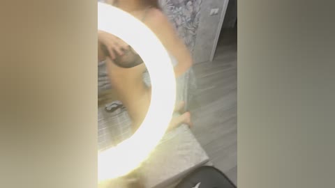 Media: A video of a nude woman with a medium build, light skin, and a tattoo on her right arm, sitting on a bed. The room has a circular mirror with a bright light, and the background includes a patterned bedspread and wooden flooring.