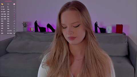 Video of a blonde woman with long hair, wearing a white top, sitting in a modern living room with gray couch, purple lighting, and black high heels on the couch.