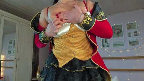 Video of a woman in a Victorian-style outfit with gold corset, black ruffled skirt, and red sleeves, adjusting her breasts, in a cozy, softly lit room with fairy lights and framed pictures on the wall.