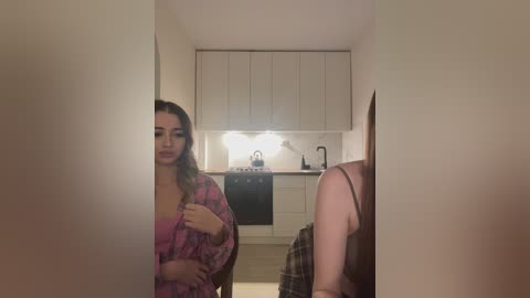 Media: Video of a young woman with light skin and long brown hair in a pink floral robe standing in a dimly lit, narrow hallway with white cabinets and a stove.