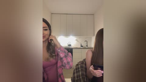 Media: Video of a woman in a pink floral shirt and black bra, adjusting her hair in a modern kitchen with white cabinets and a light countertop.