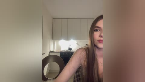 Media: Video of a young woman with long brown hair, wearing a plaid top, seated in a modern kitchen with white cabinets and a round mirror.