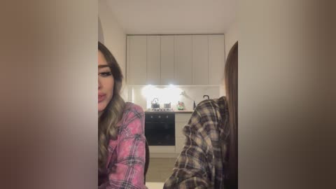 Media: Video of two young women in a narrow hallway; one with long wavy hair in a pink plaid shirt, the other in a plaid flannel. Kitchen cabinets and countertop visible in the background.