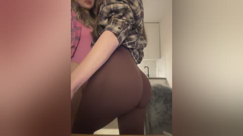 Media: A video captures a woman in a pink shirt and plaid jacket, leaning against a white wall. She wears tight, maroon leggings, highlighting her curvy buttocks. The background shows a bathroom with a sink and towel.