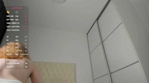 Media: A video of a nude woman with fair skin and red hair, viewed from behind, standing in a minimalist white room with a sliding door wardrobe.