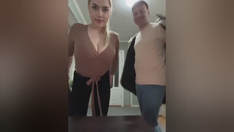 A video of a blonde woman in a low-cut, beige top and jeans, and a balding man in a white shirt, standing in a hallway with beige walls and a wooden floor.